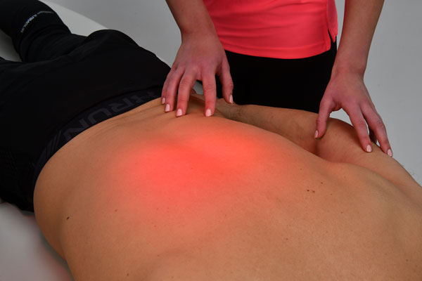 Ein Patient liegt mit freiem Oberkörper auf dem Bauch, während eine Physiotherapeutin eine Wärmeanwendung auf dem Rücken durchführt